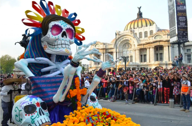 Desfile de Día de Muertos en la Ciudad de México con una catrina al lado izquierdo de la imagen, decorada con calaveras, flores de cempasúchil y ropa mexicana. Al fondo el palacio de Bellas Artes y una multitud de personas mirando el desfile.
